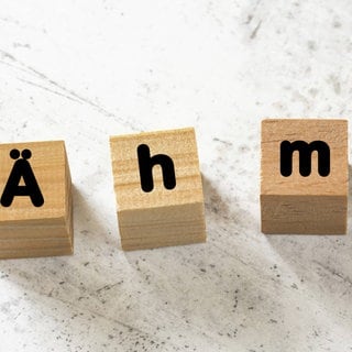 Vier Holzwürfel bilden das Füllwort "Ähm" auf einer weißen Zementplatte. Symbolfoto.
