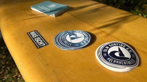 Mit Stickern auf einem Surfbrett an der Blau wirbt der Verein Ulm Surfing für seinen Traum vom Surfen auf einer Welle an der Blau.  
