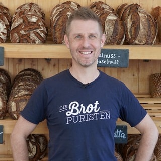 Sebastian Däuwel trägt ein T-Shirt mit der Aufschrift „Die Brotpuristen“ und steht vor einem Brotregal in seiner Bäckerei.