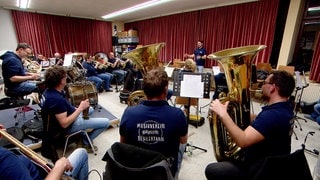 Eine Musikkapelle sitzt in einem Proberaum mit einem roten Vorhang. Im Vordergrund sitzen Menschen mit großen Tubas. Im Hintergrund ist ein Mann zu sehen, der dirigiert.