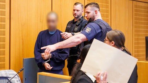 Mordprozess am Mainzer Landgericht: Der angeklagte Vater wird von Justizbeamten in den Saal geführt, die Mutter hält sich ein Papier vor das Gesicht. 