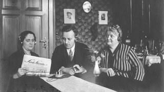 Drei Radiohörer, zwei Frauen, in der Mitte ein Mann, mit Kopfhörern und Detektorempfänger. Undatiertes Foto aus den 1920ern. Die Frau links im Bild hält ein Papier, vermutlich ein Programmheft, mit dem Titel "Funk Stunde" in der Hand.