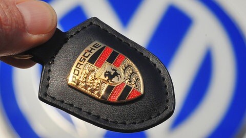 Ein Porsche-Firmenlogo wird vor ein VW-Logo in Wolfsburg gehalten