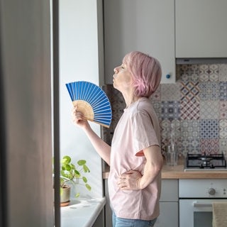 Symbolbild: Erschöpfte Frau benutzt einen Fächer, um sich bei Hitze in Innenräumen Abkühlung zu verschaffen.