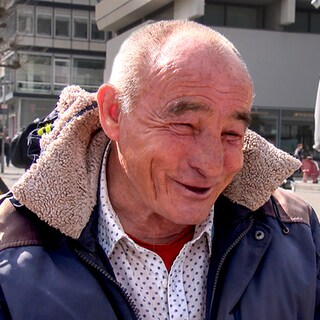 Rudolf spielt jeden Tag Schach auf dem Schlossplatz in Stuttgart. Er sitzt an seinem Klapptisch mit seinem Schachbrett und lacht in die Kamera.