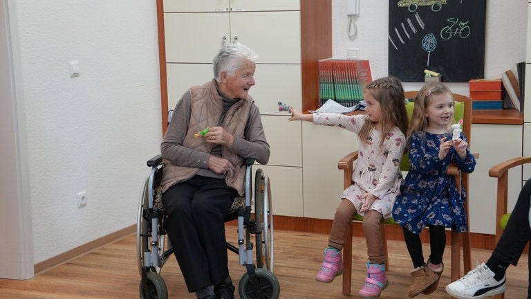 Eine ältere Frau im Rollstuhl sitzend unterhält sich mit zwei Kindergartenkindern. 