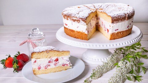 Die frische Erdbeertorte mit Biskuitboden auf einem weißen Tortenständer, ein Stück ist herausgeschnitten, rechts daneben liegt ein Holunderblütenast, links daneben ein kleiner weißer Teller mit einem herausgeschnittenen Stück Torte. Neben dem Teller liegen drei ganze Erdbeeren.