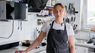 Mariia ist gelernte Köchin und Konditorin. In ihrer Küche steht sie in ihrer Schürze und blickt selbstbewusst in die Kamera. Im Hintergrund sind Schwerlastregale zu sehen, in denen sie ihre Backutensilien aufbewahrt.