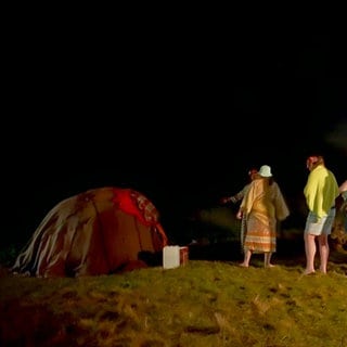 Menschen stehen im Dunkeln vor der Schwitzhütte an. Im Hintergrund brennt ein Feuer.