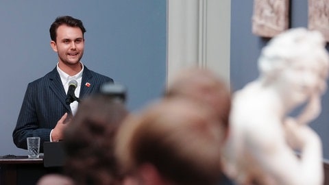 Ein Mann steht vor einer Skulptur und spricht. Es handelt sich um den Künstler Jérémie Queyras, der ein Porträt der Alt-Kanzlerin Angela Merkel gemalt hat.