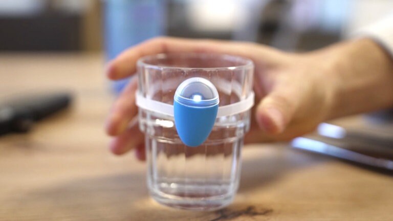 Hand stellt ein Glas mit einer blauen Glasleuchte, die um das Glas gespannt ist, auf einen Tisch ab. Die Glasleuchte soll durch blinken an das regelmäßige trinken erinnern.