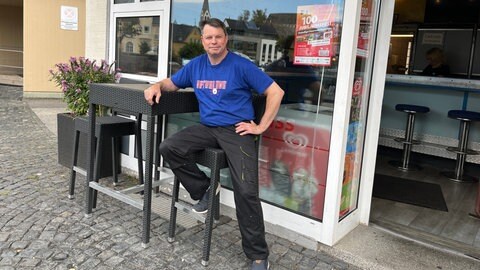 Uwe Jakobs lehnt an einem Stehtisch vor seinem Imbiss in Kastellaun. Am Wochenende werden wieder viele Stammgäste herkommen, die jedes Jahr die NATURE ONE besuchen und dann auch einen Abstecher zu ihm machen.