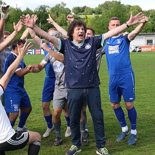 Olaf der Flipper steht mit einem dunklen Deutschland-Fußballtrikot zusammen mit Fußballern des VfB Bretten auf einem Fußballplatz. Oliver Pocher kniet vor der Mannschaft. Alle jubeln.