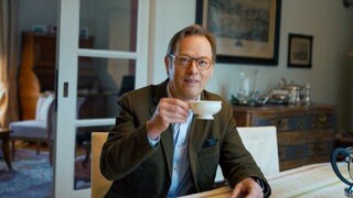 Burgherr Bernolph Freiherr von Gemmingen sitzt in eleganter Kleidung an einem Tisch in der Burg Guttenberg und hält eine Teetasse. Im Hintergrund sind historische Einrichtungsgegenstände und Bilder zu sehen.