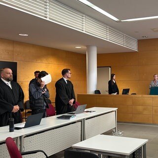 Sitzungssaal am Landgericht Kaiserslautern: Richterin, Staatsanwaltschaft, Verteidiger und Angeklagter sind versammelt. Angeklagter hält Papier vor sein Gesicht.