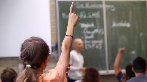 Ein Mädchen streckt im Unterricht den Finger, ein Lehrer erklärt an der Tafel: Demokratiebildung ist Aufgabe von Schule. Der sogenannte "Beutelsbacher Konsens" aus dem Jahr 1976 legt die Prinzipien dafür fest: Keine Indoktrination im Klassenzimmer, politische Kontroversen sind auch kontrovers darzustellen, zudem sollen Schüler*innen befähigt werden, sich eine eigene Meinung zu bilden. 