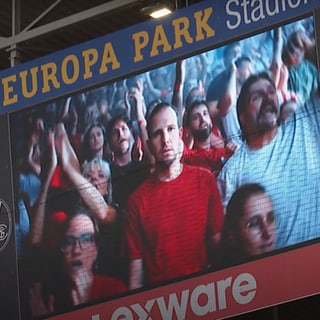 Auf der Leinwand steht ein Mann mit leerem Gesichtsausdruck inmitten von Fußballfans. 