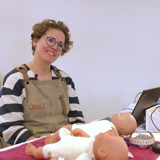 Puppenärztin Anja Müller sitzt an einem Tisch. Vor ihr liegen zwei Puppen, ein Maßband und Nähgarn. 