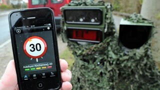 Ein Smartphone mit einer Blitzer-Warn-App wird vor eine mobile Geschwindigkeits-Messanlage gehalten. 
