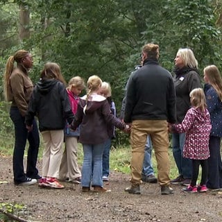 Eine Gruppe aus Erwachsenen und Kindern steht in einem Kreis im Wald.