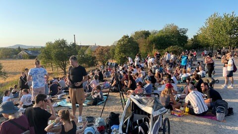 Viele Menschen sitzen auf einer Wiese am Freiburger Mundenhof. Es gibt Picknickdecken und Kinderwagen. Sie wollen die Sonnenfinsternis beobachten.