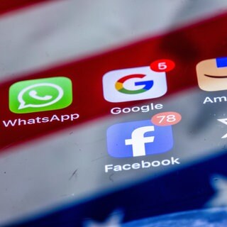 Die Apps verschiedener amerikanischer Tech-Konzerne, WhatsApp, Google, Amazon, Facebook und X, sind auf dem Display eines Smartphones zu sehen, auf dem sich die US-amerikanische Flagge spiegelt.