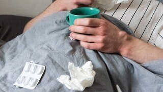 Symbolbild - ein Mann liegt im Bett und trinkt Tee und hat ein Taschentuch in der Hand
