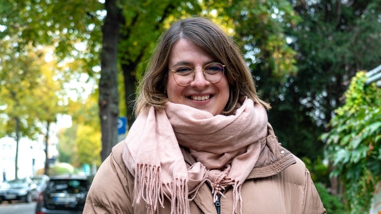 Junge Frau steht vor Bäumen und lächelt in die Kamera. 