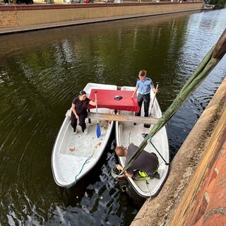 Zwischen zwei zusammengebundenen Ruderbooten hängt ein Seil ins Wasser. Im Wasser kann man den versenkten Roller erahnen.