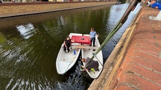 Zwischen zwei zusammengebundenen Ruderbooten hängt ein Seil ins Wasser. Im Wasser kann man den versenkten Roller erahnen.