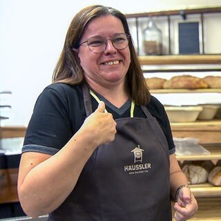 Luzia ist Kursteilnehmerin im ersten Backkurs mit Gebärdensprache. Sie steht in einer Backschürze vor einem Regal mit vielen frischen Broten. Sie hat ihre schulterlangen Haare offen, trägt eine Brille und lächelt in die Kamera.