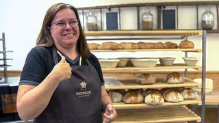 Luzia ist Kursteilnehmerin im ersten Backkurs mit Gebärdensprache. Sie steht in einer Backschürze vor einem Regal mit vielen frischen Broten. Sie hat ihre schulterlangen Haare offen, trägt eine Brille und lächelt in die Kamera.