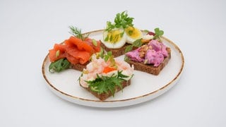 Vier verschiedene Smørrebrød auf einem Teller. Smörrebröd ist mehr als ein belegtes Brot: Es sind kleine Kunstwerke, die liebevoll angerichtet werden. Die reichlich belegten Butterbrote sind in Dänemark ein traditionelles Mittagessen. Grundlage ist gebuttertes Roggenbrot.