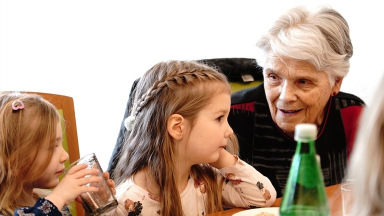 Eine ältere Frau mit kurzen grauen Haaren und schwarz-roten Pullover redet mit zwei Kindergartenkindern, die neben ihr sitzen.