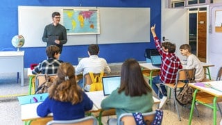 Eine Gruppe Jugendlicher hebt die Hand und folgt dem Erdkundeunterricht im Klassenzimmer mit Lehrer und Laptops.