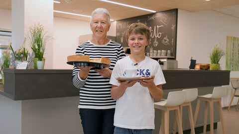 Annemarie, eine Frau in ihren 70ern und Alex, ein 11-jähriger Junge, stehen in einem Café. Annemarie hält einen Kuchen, Alex eine weiße Tasse. Die beiden strahlen in die Kamera.