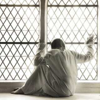 Frau in weißem Nachthemd am vergitterten Fenster einer Psychiatrie (Symbolbild): Bis in die 1970er-Jahre glichen Psychiatrien elenden Verwahranstalten: Patienten wurden ruhiggestellt, weggesperrt und unter menschenunwürdigen Bedingungen untergebracht.