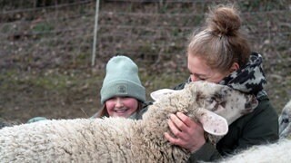 Eine Frau kuschelt mit einem Schaf. Im Hintergrund lächelt ein Kind.