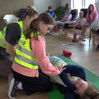 Ein Kind sitz mit einer Frau auf dem Boden. Beide tragen eine gelbe Warnweste. Vor ihnen liegt eine Frau auf dem Boden. Um sie herum sitzen weitere Kinder.