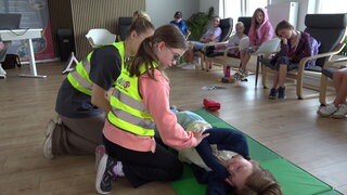 Ein Kind sitz mit einer Frau auf dem Boden. Beide tragen eine gelbe Warnweste. Vor ihnen liegt eine Frau auf dem Boden. Um sie herum sitzen weitere Kinder.