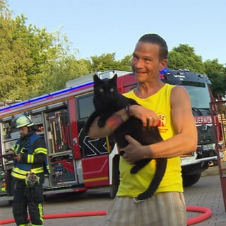 Leiter einer Tiepension hält eine Katze auf dem Arm während im Hintergrund eine Feuerwehrübung stattfindet.