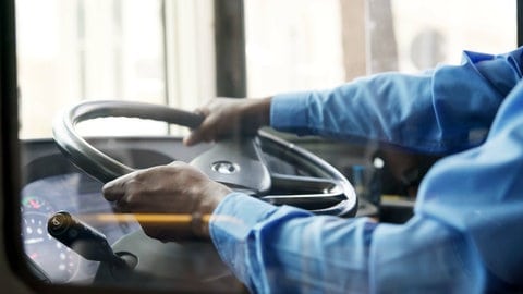Ein Busfahrer sitzt am Steuer eines Busses. Ein Lenkrad ist zu sehen.