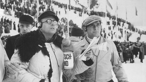 Paul Laven berichtet im Februar 1936 auf dem Olympiagelände in Garmisch-Partenkrichen über einen Abfahrtslauf, während Paul Müller (Olympiabeauftragter der Reichssendeleitung) neben ihm steht. Im Hintergrund Publikum. 