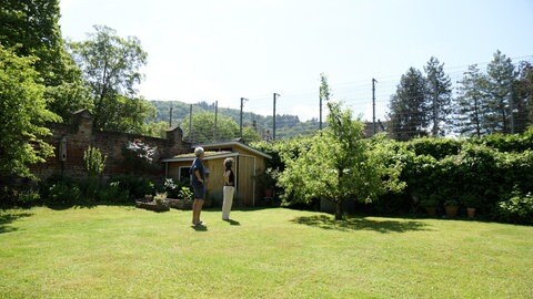 Zwei Menschen stehen in einem Garten. In der einen Ecke befindet sich eine kleine Gartenhütte. Dahinter ein Zaun, der zu einem Sportplatz gehört.