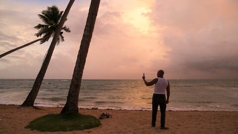 Ein Mann steht an einem Karibik-Strand und macht bei Sonnenuntergang ein Foto mit seinem Smartphone.