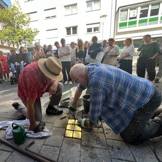 Menschen stehen auf einem Bürgersteig vor einem Haus und tragen Texte vor, währenddessen werden goldfarbene Steine im Boden verlegt. Sechs Stolpersteine werden für die Familie Hirschfeld vor dem ehemaligen Wohnhaus verlegt. 