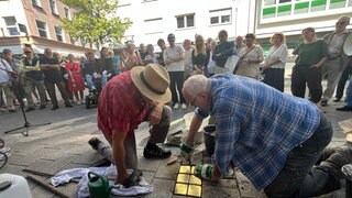 Menschen stehen auf einem Bürgersteig vor einem Haus und tragen Texte vor, währenddessen werden goldfarbene Steine im Boden verlegt. Sechs Stolpersteine werden für die Familie Hirschfeld vor dem ehemaligen Wohnhaus verlegt. 