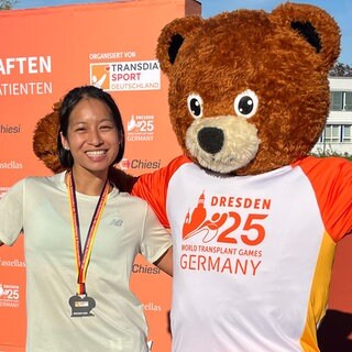 Rasa Fuchs steht links von einem Bären-Maskottchen. Sie trägt eine Medaille um den Hals und lächelt in die Kamera. Der Bär trägt ein T-Shirt mit der Aufschrift "Dresden 25 World Transplant Games Germany".