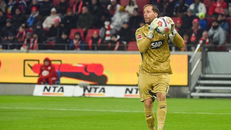 Oliver Baumann trägt ein goldenes Torwarttrikot und hält einen Fußball in der Hand