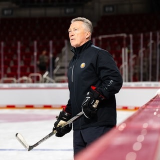 Eishockey-Nationaltrainer Harold Kreis hält einen Eishockey-Schläger in den Händen und steht auf dem Eis. 